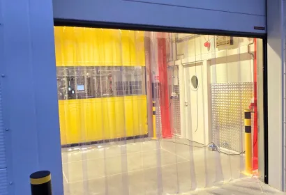 Przezroczysta kurtyna paskowa PCV w otworze bramy rolowanej, za nią żółte pasy wydzielające strefę i słupki ochronne
